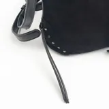 Cartera de hombro color negro, confeccionada en material tipo gamuza, con correa ajustable y detalles de tachas metálicas en las esquinas inferiores. Presenta una tira vertical central con hebilla decorativa y tiras laterales colgantes.