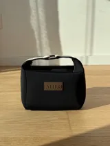 Neceser de neopreno color borgoña, con cierre superior y forro interior. Tiene un asa de mano con una franja azul y blanca.