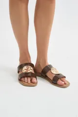 Sandalias planas color marfil con dos tiras anchas en el empeine, decoradas con un aplique metálico dorado de diseño orgánico en el centro. Suela marrón.