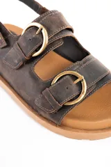 Sandalias de cuero marrón oscuro, con doble tira ancha en el empeine y tira en el talón, todas con hebillas metálicas doradas. Cuenta con suela plana y gruesa de tono tostado con estriado inferior.
