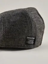 Boina estilo irlandés en color gris con textura jaspeada. Presenta un detalle lateral en cuero marrón con un botón metálico decorativo y visera integrada.