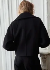 Chaqueta negra de corte corto y holgado, confeccionada con mezcla de lana. Cuenta con cuello ancho y solapa de piel sintética en tono beige claro, cierre frontal con botones oscuros y mangas largas amplias.
