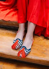 Sandalias planas destalonadas confeccionadas en rafia trenzada con patrón de zigzag en tonos azul marino y crudo. Tienen punta cerrada con una borla decorativa de flecos naranjas en el empeine, y una tira trasera ajustable de cuero naranja con hebilla metálica dorada.