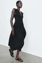 Vestido midi negro de escote pico y manga sisa, con detalles de bordados combinados a tono. Presenta un tejido plisado con efecto arrugado y cierre en la espalda con lazada y cremallera oculta.