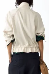 Campera corta de lino color marfil con cuello alto, solapa con botones, bolsillos diagonales con solapa, mangas raglán y cordón ajustable en la cintura.