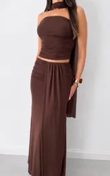 Conjunto de dos piezas marrón, compuesto por un top strapless fruncido con accesorio para el cuello y una pollera larga con drapeado frontal.