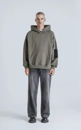 Hoodie oversize color verde militar, confeccionado en algodón rústico. Presenta capucha forrada, bolsillo frontal tipo canguro y un bolsillo adicional con cierre en la manga izquierda, con detalles en microfibra negra.