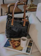 Bolso tipo tote de cuero negro con acabado brillante y detalles de costuras y asas en cuero color suela. Cuenta con bolsillo frontal externo, compartimento principal con cierre y doble asa de mano, además de correa larga ajustable.