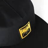 Gorra snapback negra con visera plana y parche frontal rectangular de color dorado con un diseño gráfico abstracto que simula dos caras o formas simétricas.