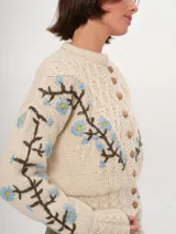 Cárdigan tejido a mano color crudo, con diseño de trenzas y flores bordadas en azul y marrón. Tiene cuello redondo y cierre frontal con botones de madera.