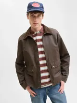 Campera de estilo worker color marrón, con cuello camisero de pana, cierre frontal con cremallera y bolsillos frontales con solapa y broches a presión. Presenta un acabado recubierto que le otorga una textura sutilmente brillante y resistente.