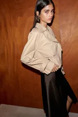 Trench corto de color beige con cuello de solapa y escote en pico. Presenta un diseño de manga larga con trabillas en los puños, bolsillos laterales y un bajo con acabado abullonado gracias a un elástico interior. Cuenta con cierre frontal cruzado con botones y forro interior.
