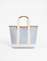 Bolso tipo tote bag de tamaño mediano, color celeste claro, con detalles en color cuero (marrón) en el borde superior y asas cortas. Presenta una cinta tejida de color crudo que recorre la base y sube por los laterales.