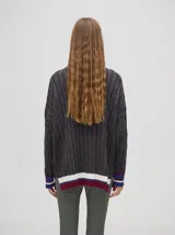 Pullover de algodón color beige, con tejido en ochos, cuello polo con cierre frontal y puños a rayas de colores (blanco, bordó y amarillo). El diseño es holgado y cae por debajo de la cadera.