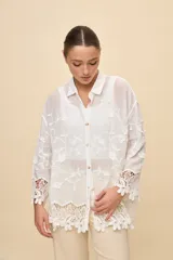 Camisa blanca de calce relajado, con cuello clásico, mangas 3/4 y cierre frontal de botones. Presenta bordados florales en relieve y detalles de crochet calado en los puños y el ruedo.