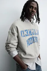 Sudadera gris de corte boxy fit, con cuello redondo y manga larga. Presenta un estampado frontal con texto en azul con efecto desgastado y acabados en rib en puños y ruedo.