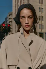 Trench largo de color negro con cuello de solapa, cierre frontal cruzado con botones y cinturón ajustable a tono. Presenta mangas largas con trabillas en los puños, bolsillos delanteros de vivo y abertura en la espalda.