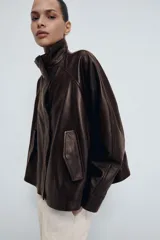 Chaqueta bomber de piel color marrón oscuro, con cuello alto, cierre frontal con cremallera y solapa con botones ocultos. Presenta mangas largas, bolsillos delanteros y un diseño de corte recto.