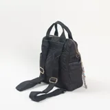 Mochila negra con diseño acolchado y pespuntes geométricos. Cuenta con un compartimento principal con cierre, un bolsillo frontal con cierre, asas superiores de mano y correas ajustables. Incluye un colgante decorativo con cuentas de colores en el lateral.
