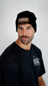 Gorro tipo beanie de punto color negro con parche rectangular bordado en el frente que incluye el logo de la marca Duxon en blanco y naranja.