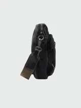 Bandolera pequeña de tela denim negra con efecto desgastado, con múltiples bolsillos frontales y cierre superior de cremallera. Incluye correa ajustable de tela tejida en tono verde oliva/marrón.