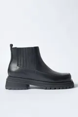 Bota corta tipo Chelsea de cuero negro, con puntera cuadrada y suela track gruesa y dentada. Presenta paneles elásticos acanalados a los lados para facilitar el calce.