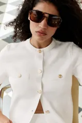 Chaqueta de punto color amarillo pálido, con cuello redondo, manga corta, cierre frontal con botones dorados y bolsillos de parche en el pecho.