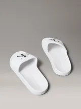 Ojotas tipo slide Calvin Klein Jeans, color blanco, con banda superior de lona de algodón que presenta el monograma de la marca en color negro. Posee suela de EVA con plantilla ergonómica moldeada.