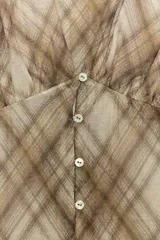 Camisa de manga larga con escote en V y cierre frontal con botones. Presenta un estampado de cuadros en tonos beige y marrón, con detalles de pliegues en el delantero y puños ajustados.