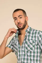 Camisa de manga corta con estampado de cuadros en tonos verdes, blancos y amarillos. Presenta cuello clásico, cierre frontal con botones y dos bolsillos delanteros con solapa en el pecho.