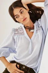 Camisa de algodón color celeste, con cuello mao, cierre frontal parcial con botones y detalles de encaje en los hombros y canesú. Presenta mangas largas abullonadas con puños elásticos.