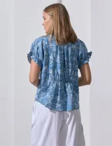 Blusa de lyocell color celeste con estampado de motivos playeros en blanco. Tiene cuello a la base con escote en V, mangas cortas con volados y detalle fruncido en mangas y espalda.