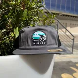 Gorro tipo trucker de la marca Hurley, color gris oscuro con malla trasera negra. Presenta un parche frontal con el logo de Hurley y un diseño de olas en tonos turquesa, blanco y gris.
