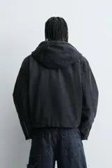 Cazadora tipo bomber de denim color negro con efecto lavado, corte boxy fit, cierre frontal de cremallera, capucha ajustable con cordón y bolsillos laterales de vivo.