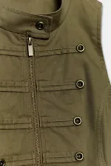 Chaleco verde militar entallado, de cuello subido y manga sisa, con detalle de aplicación de botones frontales y cierre con cremallera.