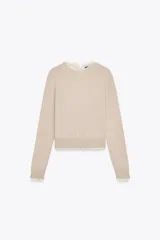 Jersey de punto color beige claro, manga larga y cuello redondo. Presenta un detalle de tejido a contraste (posiblemente una tela blanca con estampado sutil) asomando en el ruedo y el cuello.