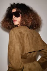 Trench corto de efecto ante color marrón, con cuello solapa, cierre cruzado con botones y cinturón ajustable del mismo tejido. Presenta mangas largas con trabillas en los puños y bolsillos delanteros de vivo.