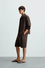 Bermuda de corte regular fit, color marrón oscuro, confeccionada en tejido con lino. Presenta pliegues frontales, bolsillos delanteros y bolsillos traseros de vivo. Cierre frontal con cremallera y botón.