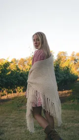 Ruana tejida a mano en forma triangular, color crudo, confeccionada en lana merino con detalle de flecos largos en los bordes.