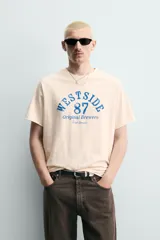 Camiseta relaxed fit de algodón color beige claro, con cuello redondo y mangas raglán cortas. Cuenta con estampado frontal en tono azul con efecto desgastado que incluye texto y el número 87.