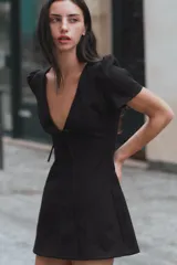 Vestido mini negro con escote en V amplio, mangas cortas abullonadas y detalle de lazo en el delantero. Cuenta con cierre de cremallera oculta en la espalda.