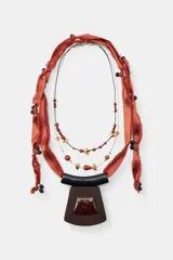 Pack de dos collares de diseño artesanal. El primero presenta una cinta de satén color terracota con flecos, adornada con cuentas negras y un colgante central de madera oscura con una pieza de resina incrustada. El segundo es un collar doble compuesto por una cadena metálica fina y un cordón negro, decorado con cuentas de piedra natural en tonos rojos, marrones y piezas metálicas doradas.