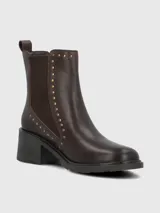 Bota tipo botín de cuero color marrón oscuro, con elásticos laterales y detalles de tachas metálicas doradas a lo largo de la caña. Cuenta con suela gruesa y taco cuadrado de altura media.
