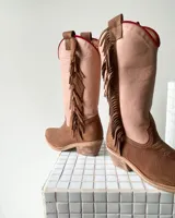 Botas estilo tejano de caña alta, confeccionadas en cuero vacuno. Presentan una combinación de colores en beige y marrón texturizado, con detalle de flecos laterales y borde superior en color rojo.