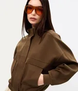 Campera marrón estilo bomber, confeccionada en punto roma, con cuello alto, mangas largas, cierre frontal oculto, bolsillos frontales aplicados y recortes en el pecho.