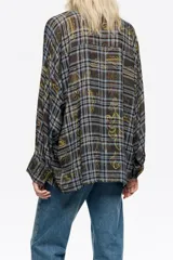 Camisa de corte oversize con cuello camisero, cierre frontal de botones y mangas largas con puños abotonados. Presenta un diseño estampado que combina un patrón de cuadros con motivos decorativos en tonos marrones, azules y amarillos.
