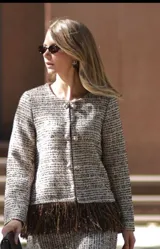 Chaqueta de tweed en tonos beige y marrón, con cuello redondo, cierre frontal con botones joya y detalle de flecos largos en el ruedo.