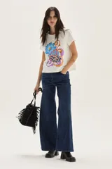 Remera de algodón color blanco, manga corta, con un estampado central que incluye una flor azul y formas abstractas con patrón a cuadros en tonos naranjas y azules, con texto bordado.