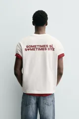 Camiseta blanca de corte relajado (relaxed fit) con cuello redondo y manga corta. Presenta un bolsillo plastrón en el pecho y un estampado de texto en color marrón rojizo en la espalda que dice "SOMETIMES SÍ, SOMETIMES BYE" (reemplazando la 'O' de 'SOMETIMES' por un corazón).
