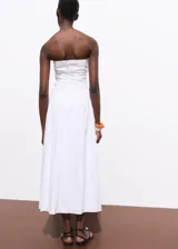 Vestido midi color blanco con diseño evasé, escote palabra de honor y sin mangas. Cuenta con detalles fruncidos en los laterales y cierre de cremallera en la parte posterior.